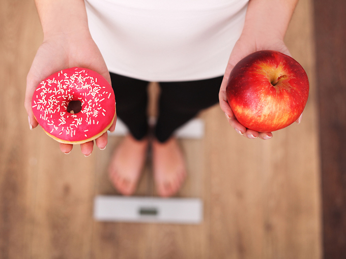 りんごとドーナツを手に体重計に乗る女性