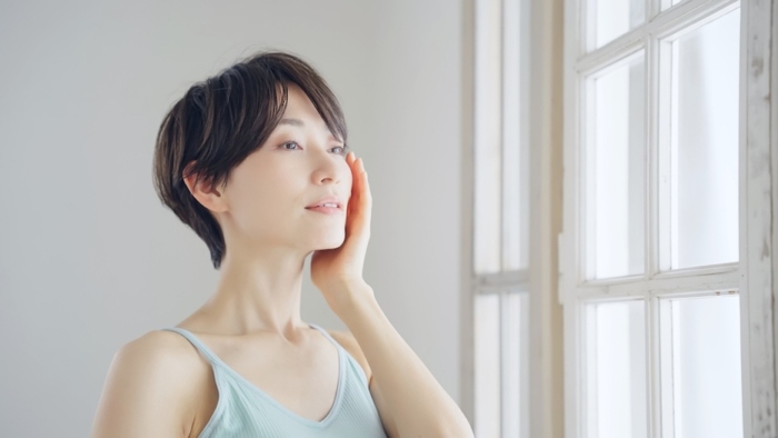 窓際で遠くを見ながら肌に手を当てる女性